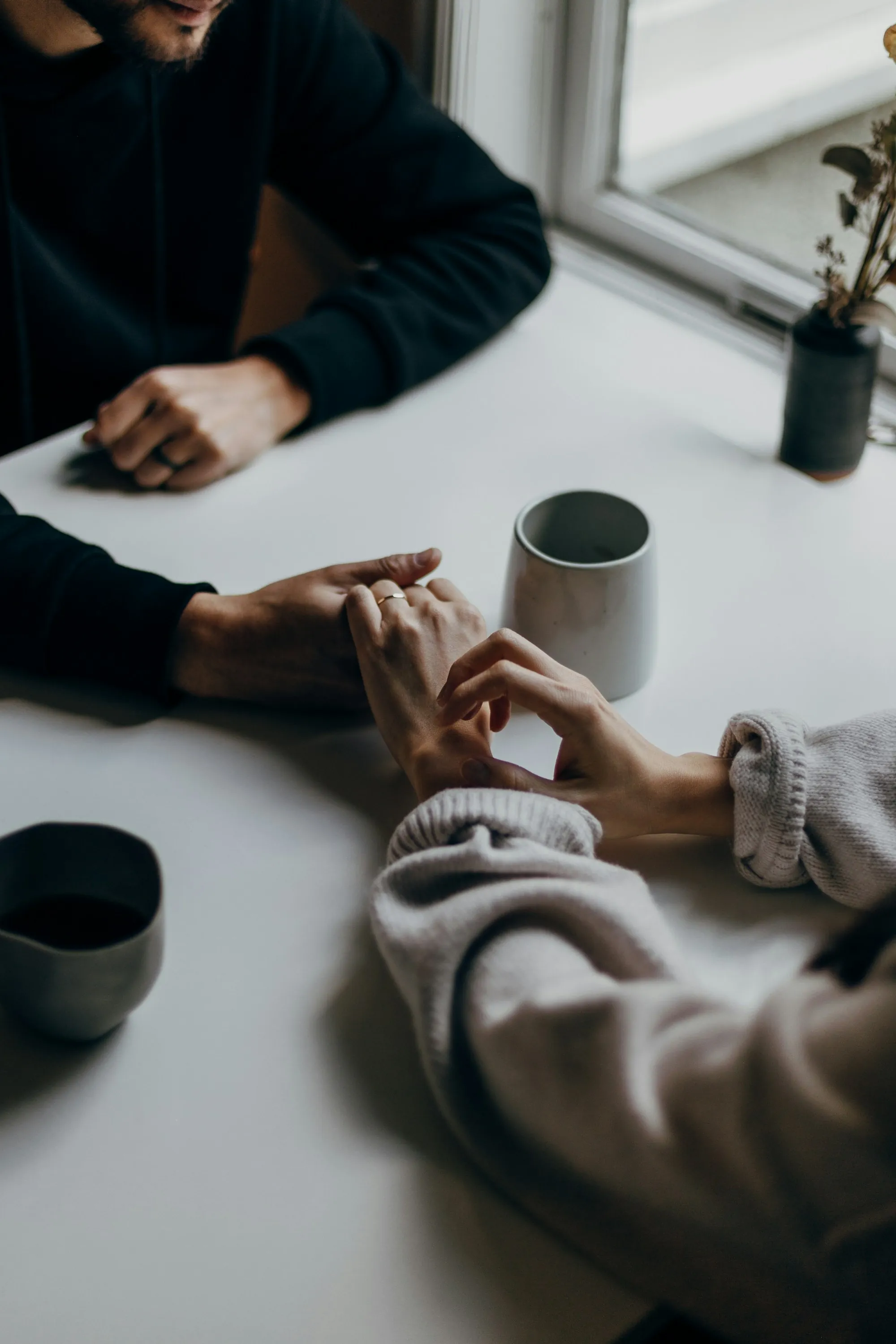 Two people sitting across a table, one holding the other's hands, warm daylight from a nearby window